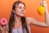 Woman in sports bra juggling choice between a donut and bell pepper on orange background.