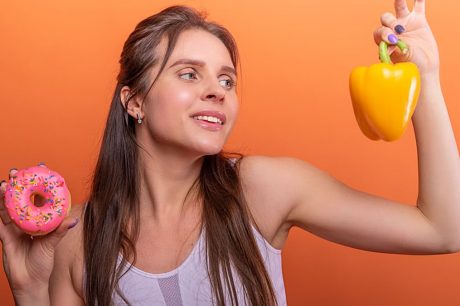 Woman in sports bra juggling choice between a donut and bell pepper on orange background.